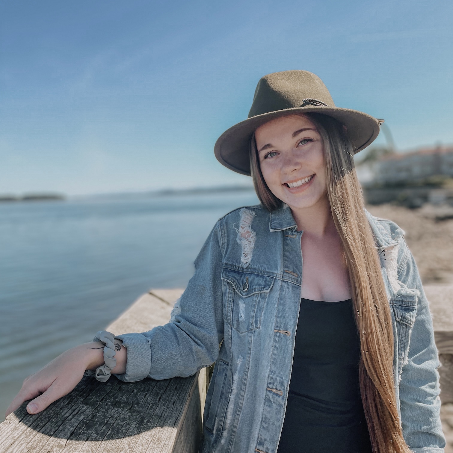 Willow McPeak smiles with her head slightly tilted and her arm resting on a wooden railing overlooking the ocean, wearing a dark tan wide-brimmed hat and denim jacket.