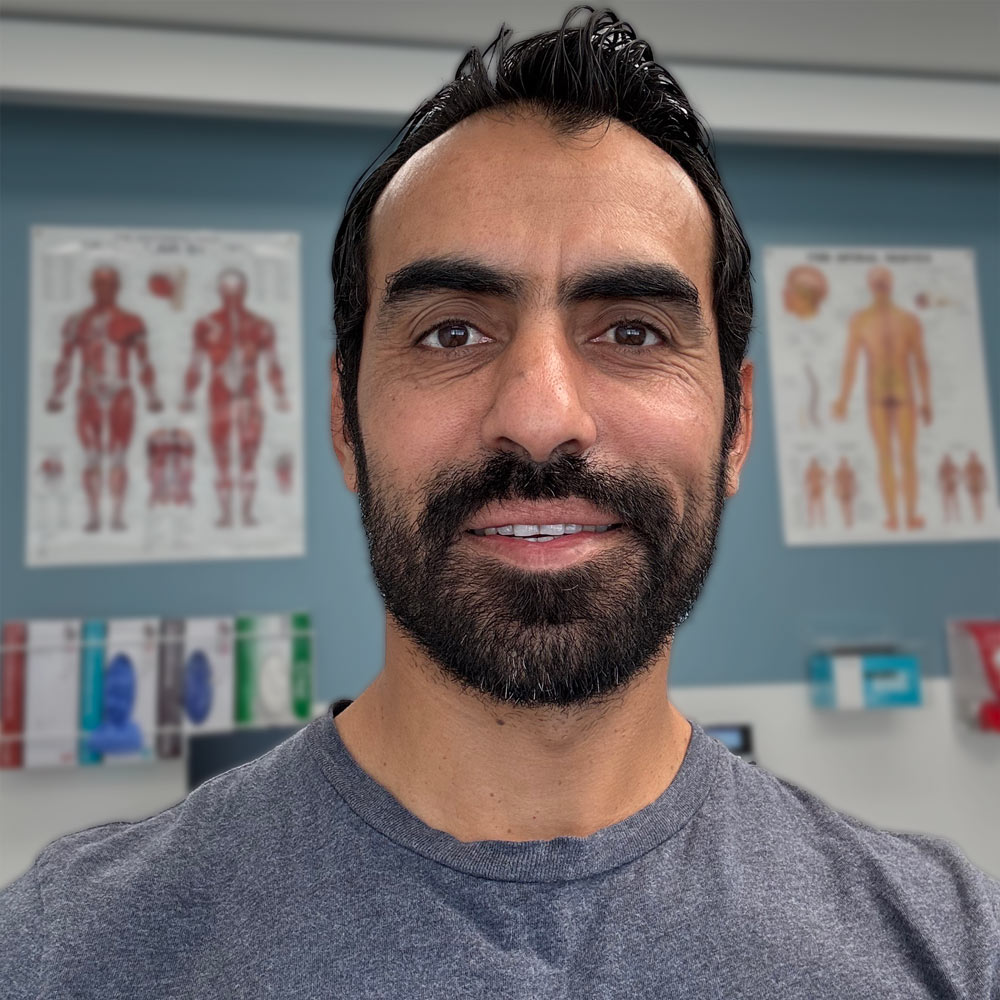 Ramze "Ramzi" Hasan, smiles widely standing in the physio clinic with anatomy posters behind him.