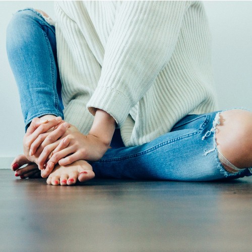 A white woman sits with one leg bent and her hands wrapped around her ankle, wearing ripped jeans and a white knit sweater.